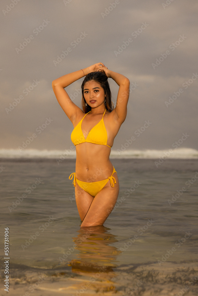 Fototapeta premium Sunset time on the beach. Sexy Asian woman in yellow bikini standing in water. Tanned skin. Long hair. Slim fit body. Tropical island vacation. Lifestyle concept. Suluban beach, Bali