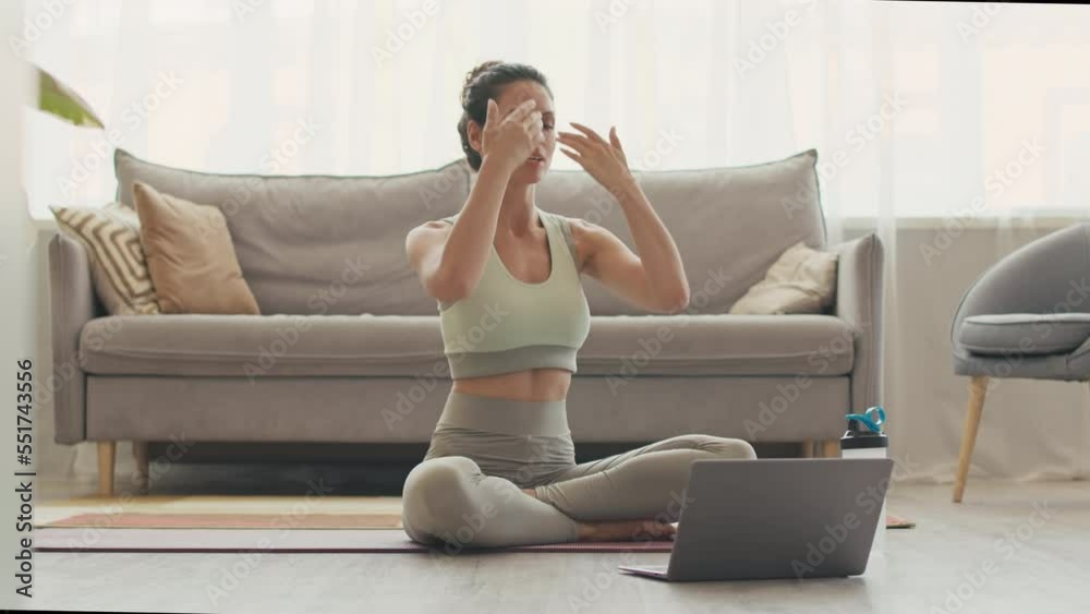 Young female yoga coach giving online video lesson via laptop, sitting on floor at home in lotus pose Young female yoga coach giving online video lesson via laptop, sitting on floor at home in lotus pose