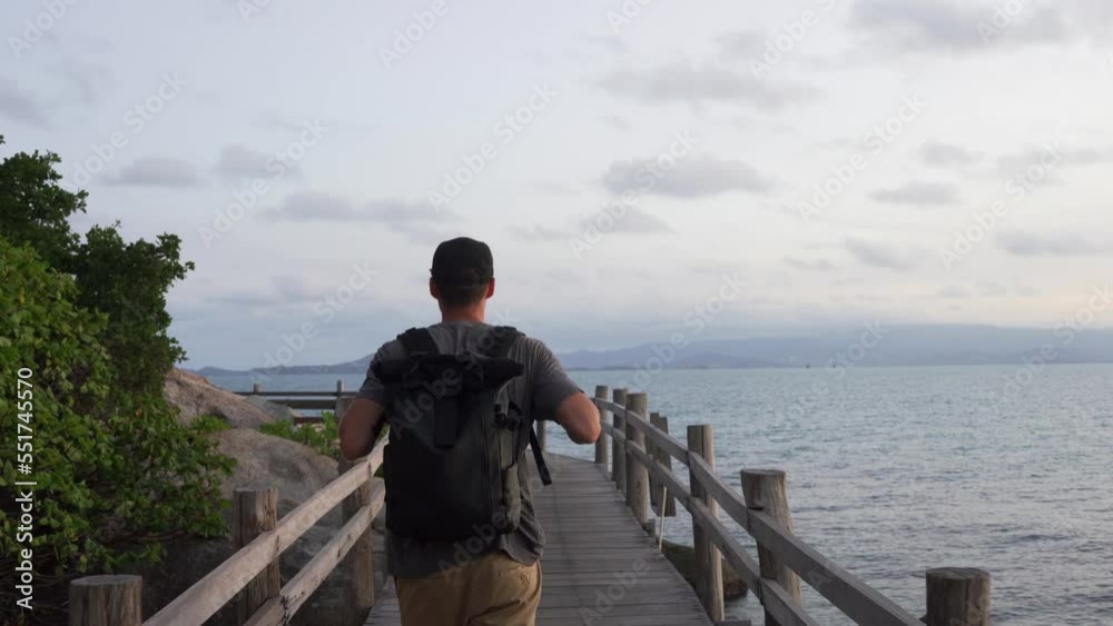 Man walking on wooden pier in tropical country near blue water. Caucasian male with backpack, walking on the dock on sunset.