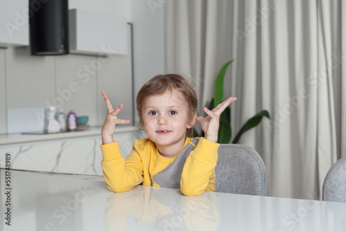 Portrait of cute child boy in yellow sweater sitting and raising his hands up on white kitchen background