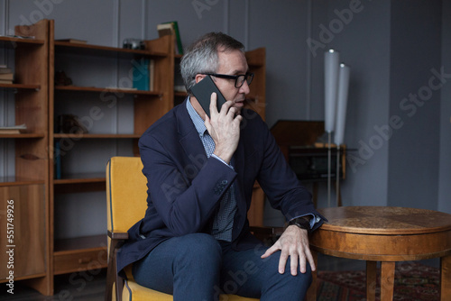 Portrait of confident serious elderly grey haired businessman in suit and glasses talking about business deals with partners on his mobile phone while sitting in chair