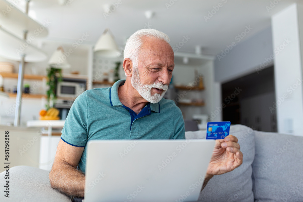 Fototapeta premium Mature man using laptop, holding plastic credit or debit card, senior grey haired customer making secure internet payment, shopping or browsing online banking service, entering information