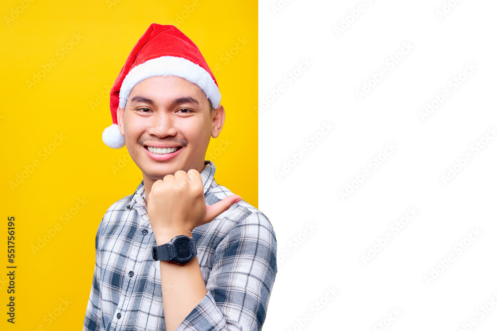 Cheerful young Asian man wearing a hat Christmas, standing and pointing thumb out the side of a white advertisement board isolated on yellow background. Happy New Year 2023 holiday concept Cheerful young Asian man wearing a hat Christmas, standing and pointing thumb out the side of a white advertisement board isolated on yellow background. Happy New Year 2023 holiday concept