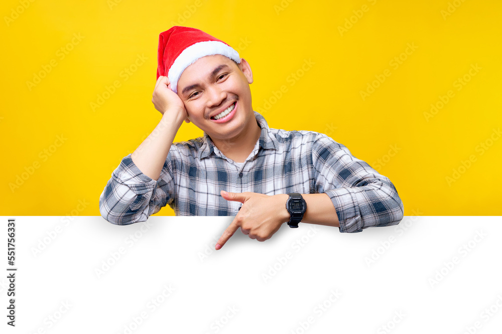 Smiling young Asian man wearing a hat Christmas pointing down at a white advertisement board isolated on yellow background. Happy New Year 2023 holiday concept