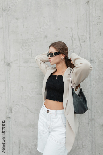 Female street portrait of a beautiful fashionable woman with stylish sunglasses in a fashion blazer, top and jeans white shorts with a handbag near the concrete wall in the city
