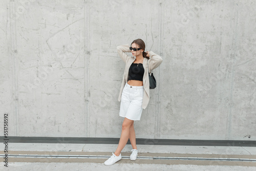 Fashion beautiful girl with black sunglasses in stylish casual clothes with a blazer, top and shorts with sneakers and a bag near a gray concrete wall