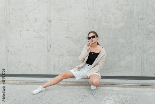 Stylish beautiful fashionable hipster girl with sunglasses in fashion casual clothes with blazer, top, shorts and sneakers sits near a gray concrete wall on the street
