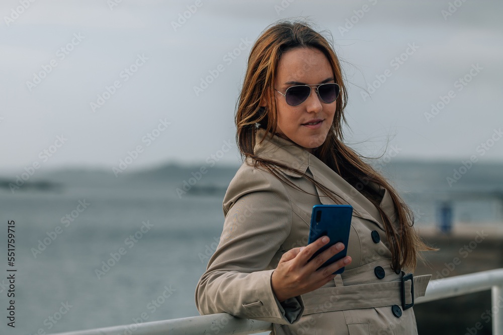 young woman in trench coat with mobile phone in port