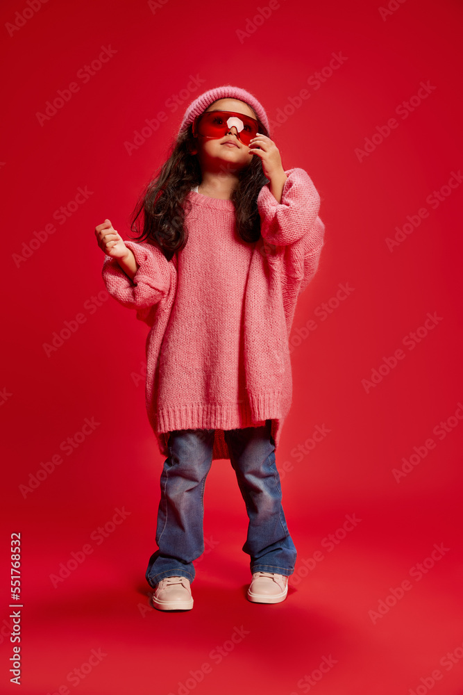 Little girl, child, posing in oversized pink sweater, red glasses and knitted hat isolated over red background. Casual. Concept of childhood, fashion, emotions Little girl, child, posing in oversized pink sweater, red glasses and knitted hat isolated over red background. Casual. Concept of childhood, fashion, emotions