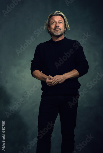 Expressive blond man with stubble beard in black sweater and jeans stands in front of a gey wall.