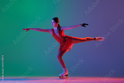 Young sportive girl, junior female figure skater in red stage costume skating isolated over gradient green-blue background in neon light. Grace, beauty, winter sports photo