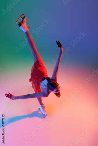 Aerial view of sportive teen girl, junior female figure skater in red stage costume skating isolated over gradient green-blue background in neon light. Grace, beauty, winter sports