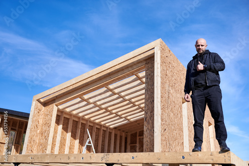 Developer building wooden frame house in Scandinavian style barnhouse. Bald man standing on construction site, inspecting quality of work, showing thumbs up on sunny day with blue sky on background. © anatoliy_gleb