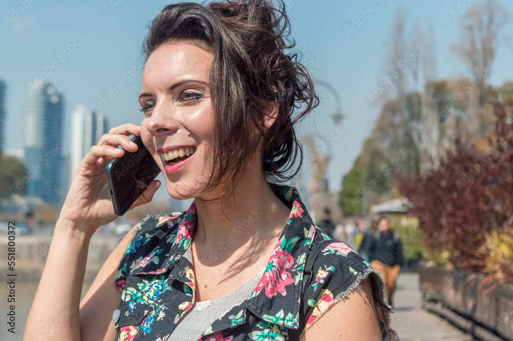 adult woman is smiling and talking on the phone outdoors.