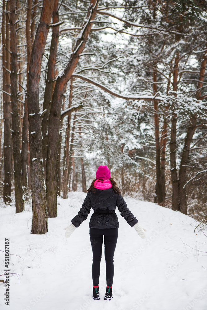 A girl in a pink scarf and hat stands with her back in the middle of a pine forest. Beautiful pine forest on a frosty day.