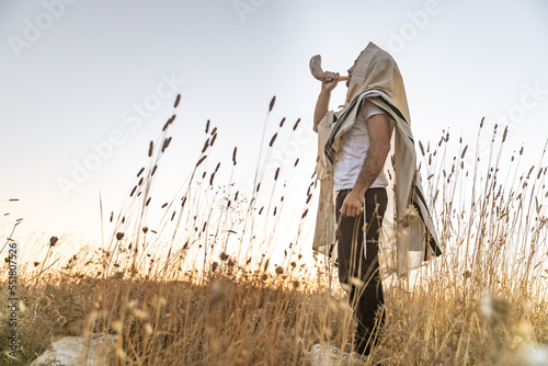 Jewish man in a traditional tallit prayer shawl blowing the ram's horn shofar, in the field against sunrise sky on Rosh HaShana and Yom Kippurim 
