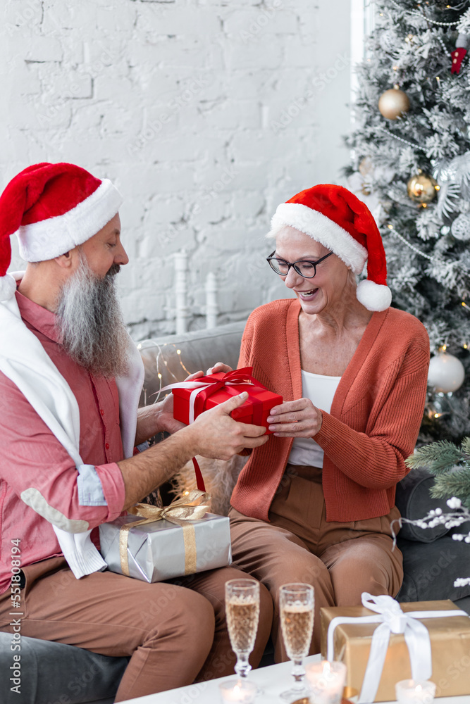 Fototapeta premium Happy loving senior couple having fun on New Year party near Christmas tree. Mature man and woman smiling, drinking champaign, exchanging gifts. Concept of active life and leisure of pensioner