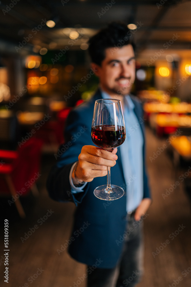 Vertical selective shot of positive elegant man in fashion suit holding glasses of red wine standing in restaurant, smiling looking at camera. Bearded male guest resting, having dinner alone.