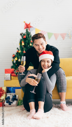 Young happy Asian surprises and laughs in the back at his girlfriend wearing a Santa Claus hat while drinking champagne at home with a Christmas tree in the background. Vertical Image.