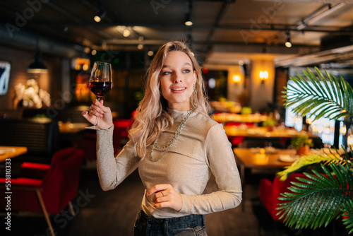 Portrait of pretty blonde lady holding glass of red wine standing in restaurant with luxury dark interior, smiling looking away. Front view of cute woman posing at cafe, blurred background.