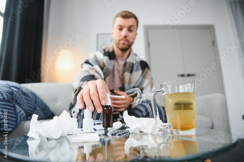 Sick bearded man who has bad cold or seasonal flu sitting on couch at home. Guy with fever wearing warm plaid shivering with worried face expression.