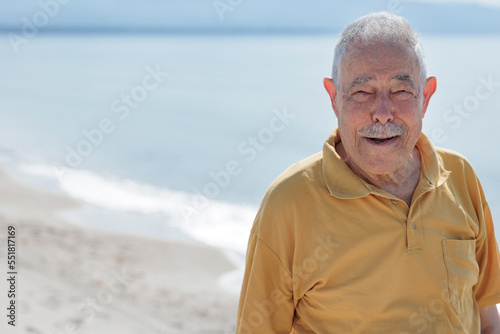 Un signore anziano vestito con polo color senape guarda sorridente in riva al mare in una spiaggia