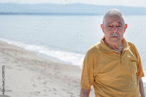 Un signore anziano vestito con polo color senape osserva serio in riva al mare in un apisaggia