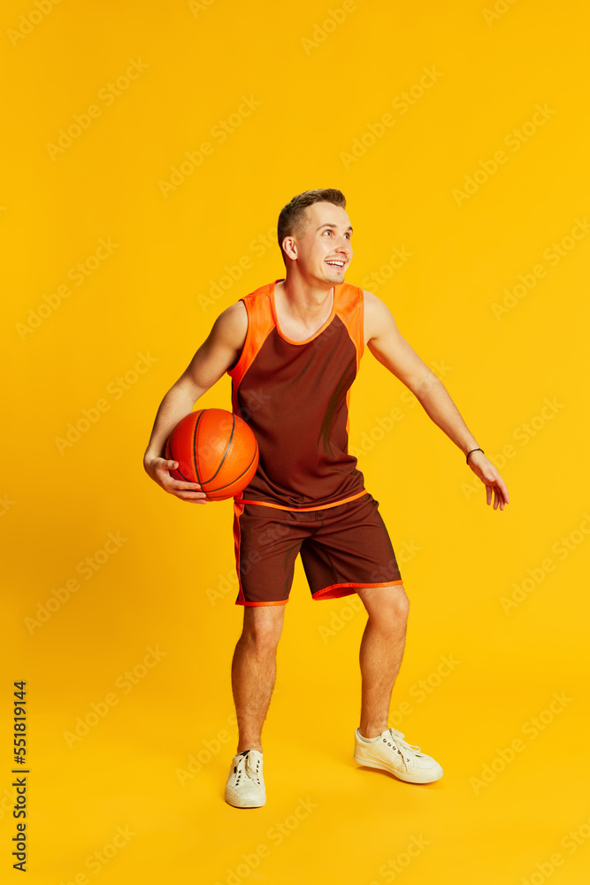 Portrait of young active man in orange uniform training, playing basketball isolated over yellow background. Fave game. Concept of sport and emotions