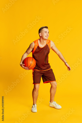 Portrait of young active man in orange uniform training, playing basketball isolated over yellow background. Fave game. Concept of sport and emotions © master1305