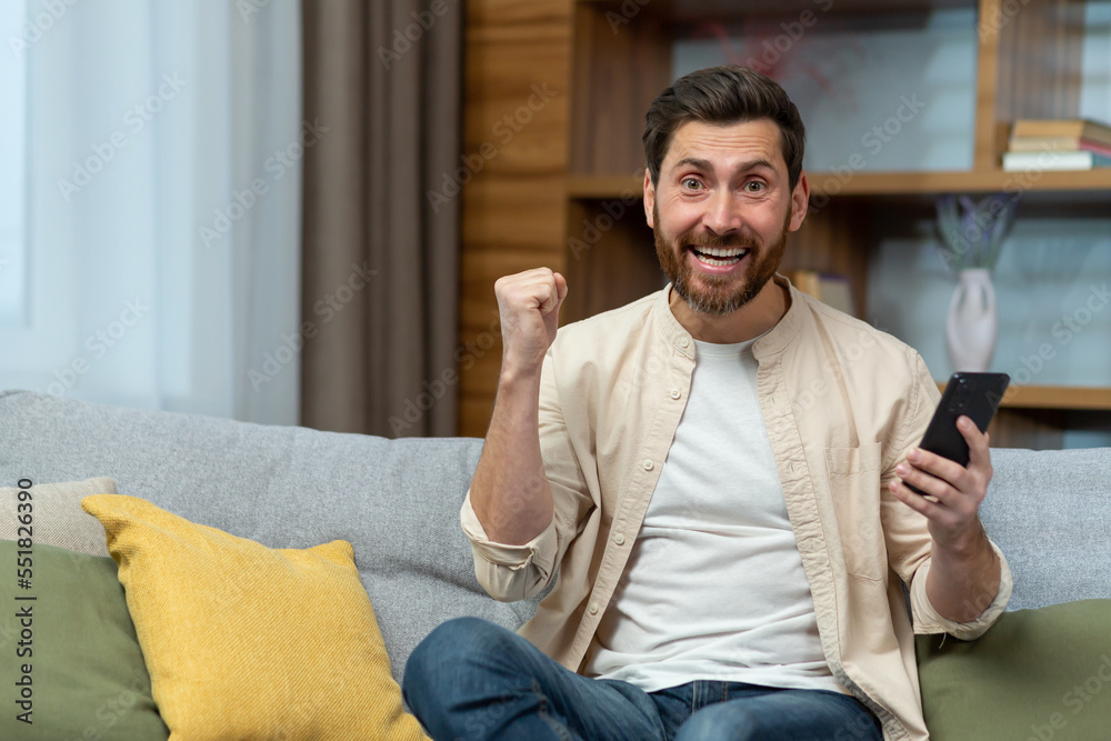 Portrait of man at home happy and celebrating online casino victory, rejoices satisfied with achievement and looks at camera holding smartphone and hand raised in triumph gesture.