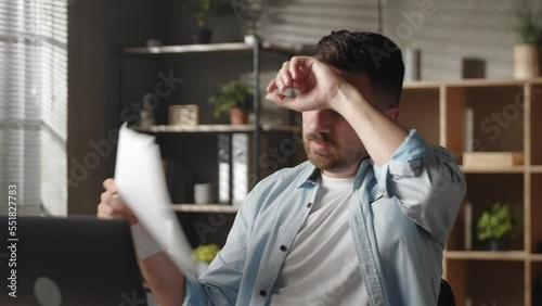 Unhappy young man sitting in hot workplace and using paper for fan