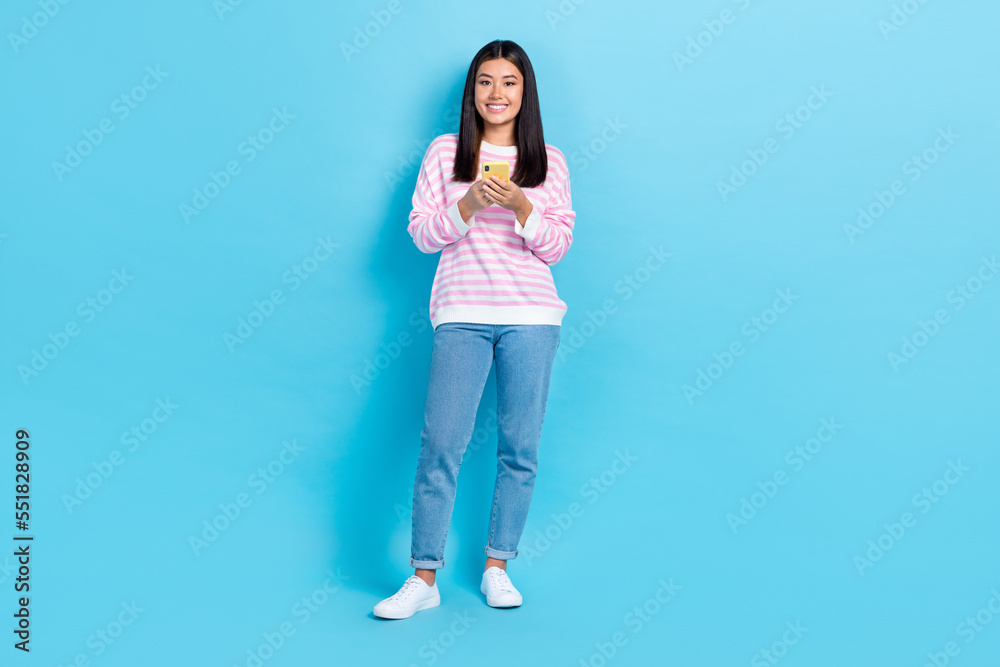 Full body length photo of young chinese girl wear casual stylish outfit hold phone chatting with her boyfriend isolated on blue color background Full body length photo of young chinese girl wear casual stylish outfit hold phone chatting with her boyfriend isolated on blue color background