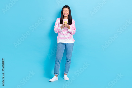 Full body length photo of young chinese girl wear casual stylish outfit hold phone chatting with her boyfriend isolated on blue color background