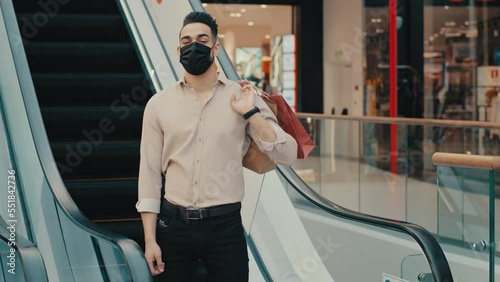 Indian Hispanic fashionable ethnic businessman trendy male shopper consumer client man buyer in face covid medical mask holding shopping bags purchases in shop store mall near escalator moving stairs photo