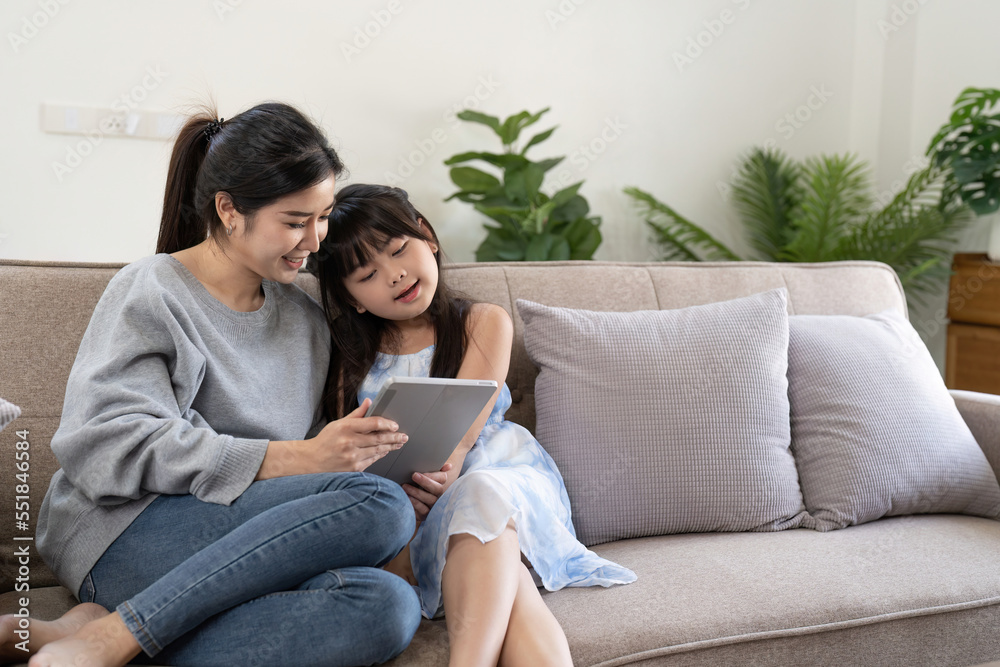 happy asian woman showing digital tablet to daughter embracing her at home. sitting on sofa