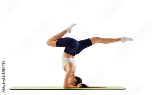 Portrait of young sportive woman training, doing balance exercises, standing on head with raised legs isolated over white background. Concept of sport, fitness, health