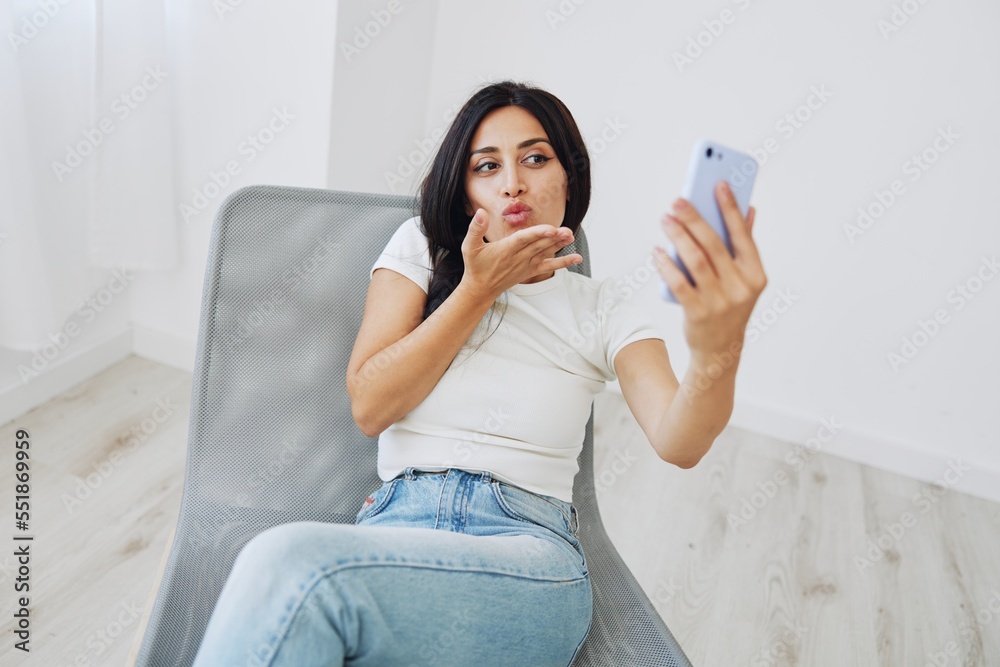 A woman with a phone sits on a chair in jeans and a T-shirt and takes pictures of herself on her phone camera, a blogger poses and takes selfies