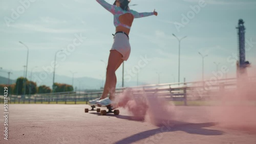 happy carefree woman is skating on longboard with pink colored smoke bomb, back view, slow motion photo