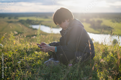 A teenage boy is sitting on the grass on a mountain against the backdrop of a beautiful sunset sky and using a mobile phone.