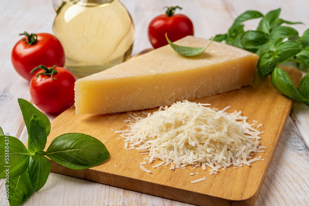 Parmesan and tomatoes on a cutting board. Italian grana padano cheese wedge and grated, red tomatoes, green basil herb and olive oil over white wooden table. Delicious hard cheese. Parmesan and tomatoes on a cutting board. Italian grana padano cheese wedge and grated, red tomatoes, green basil herb and olive oil over white wooden table. Delicious hard cheese.