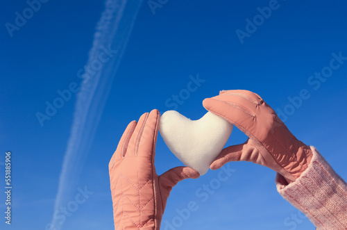 a heart made of snow in women's hands, dressed in pink gloves, against a blue sky. Selective focus