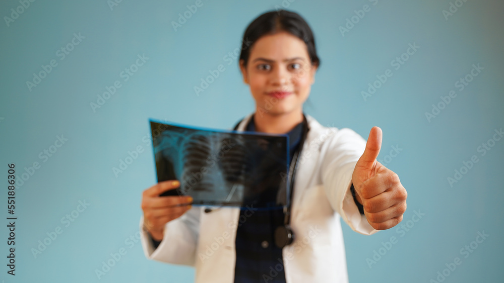Portrait of a young female doctor examining patient's x-rays scan, Cheerful Asian Indian woman doctor in apron and stethoscope isolated over blue studio background Portrait of a young female doctor examining patient's x-rays scan, Cheerful Asian Indian woman doctor in apron and stethoscope isolated over blue studio background