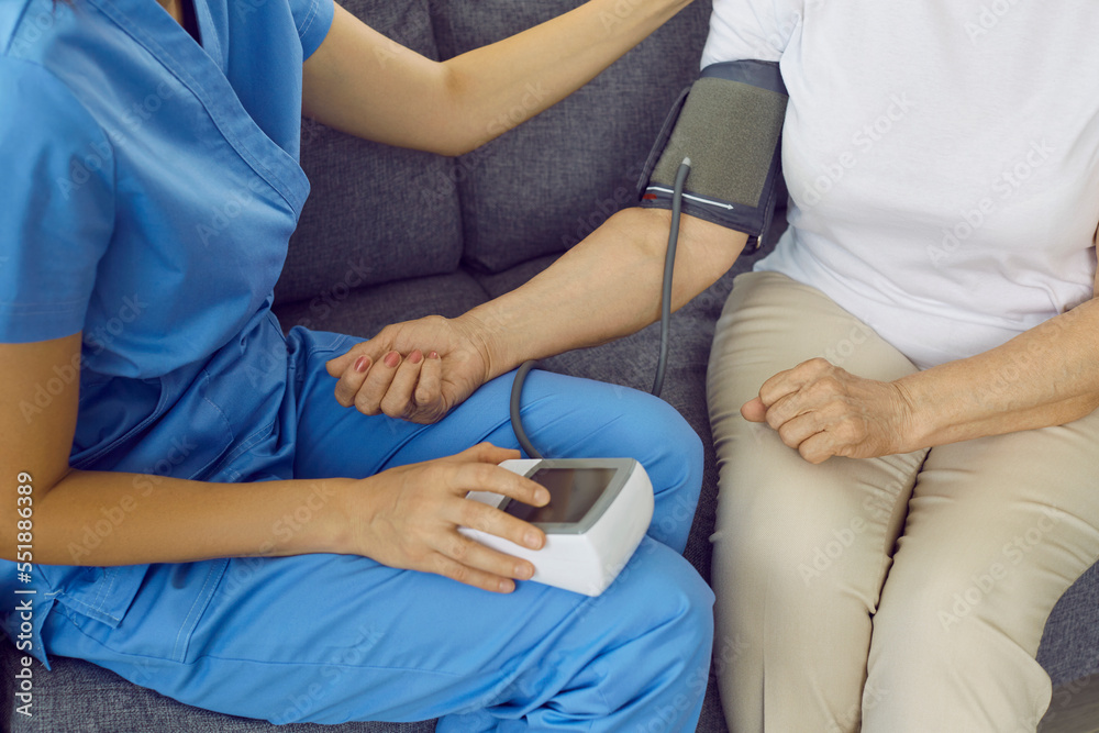 Nurse or doctor examining senior patient with hypertension. Medical specialist in blue scrubs sitting on sofa with old woman and measuring her blood pressure with tonometer. Senior health concept