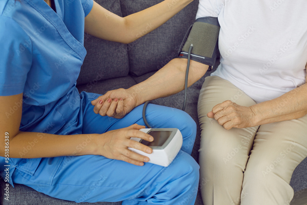 Nurse or doctor examining senior patient with hypertension. Medical specialist in blue scrubs sitting on sofa with old woman and measuring her blood pressure with tonometer. Senior health concept Nurse or doctor examining senior patient with hypertension. Medical specialist in blue scrubs sitting on sofa with old woman and measuring her blood pressure with tonometer. Senior health concept