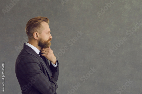 Side view portrait handsome bearded man in jacket looking hand on chin at text copyspace on grey background. Experienced business finance professional thinking about interesting quote or project idea