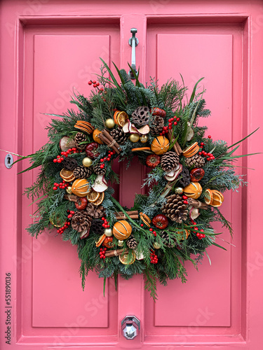 Christmas mood: festive elegant charming christmasy themed winter natural wreath on a pink wooden door. Wreath decorated with oranges, lotus, flowers snd cinnamon sticks