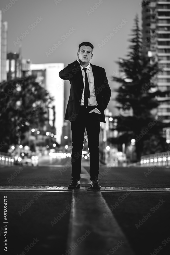 Obraz premium Young and attractive model guy with black suit on the middle of the street in the city downtown during sunset (in black and white)