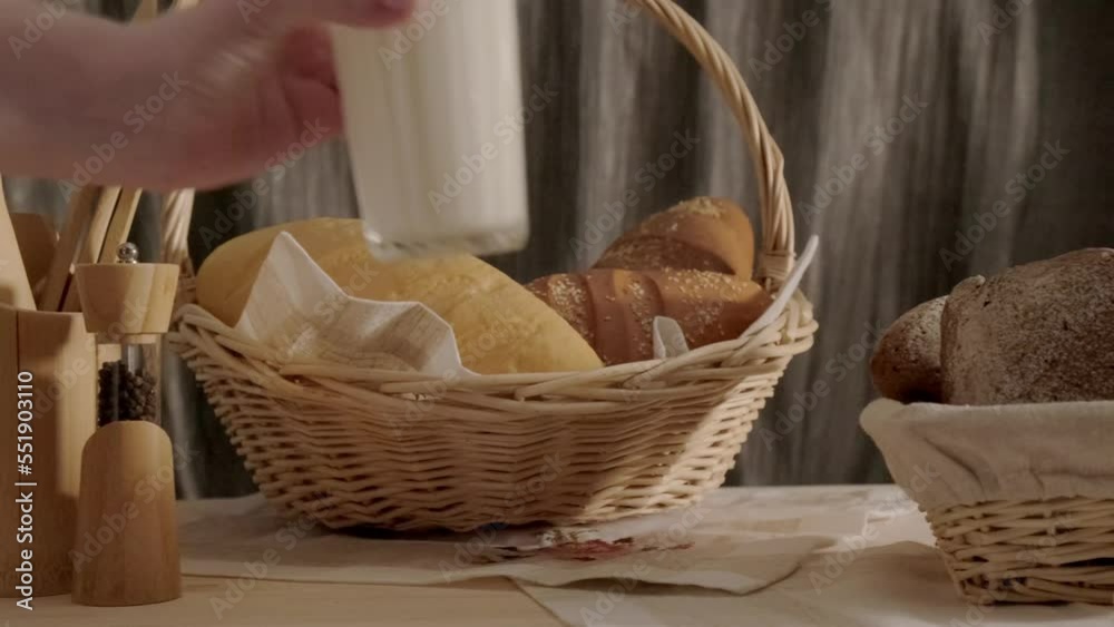 fresh baked buns of home made bread in basket and hand putting glass of milk to wooden table, bakery shop breakfast