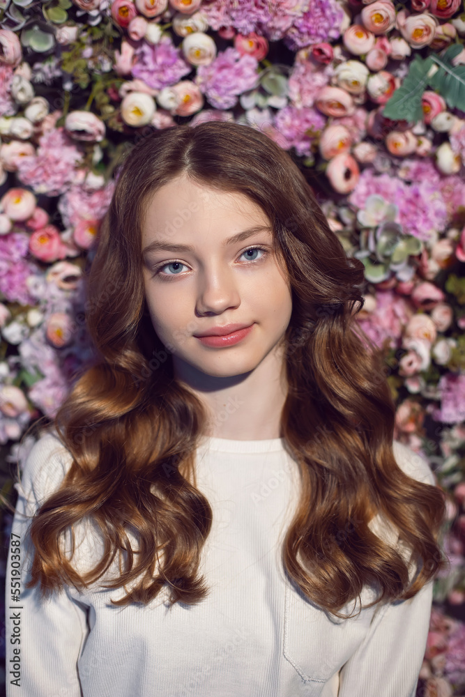 portrait of a teenage girl with long hair in a white sweater against a wall of flowers.