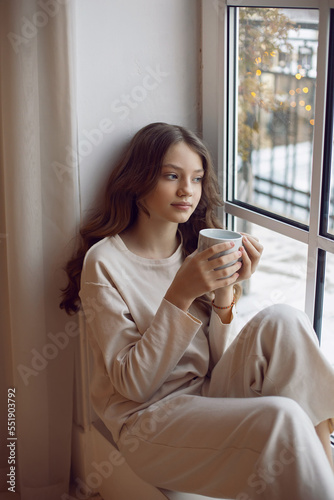 Portrait of a lonely sad teenage girl in a white warm suit sitting on the window with a cup of warm tea in winter at Christmas